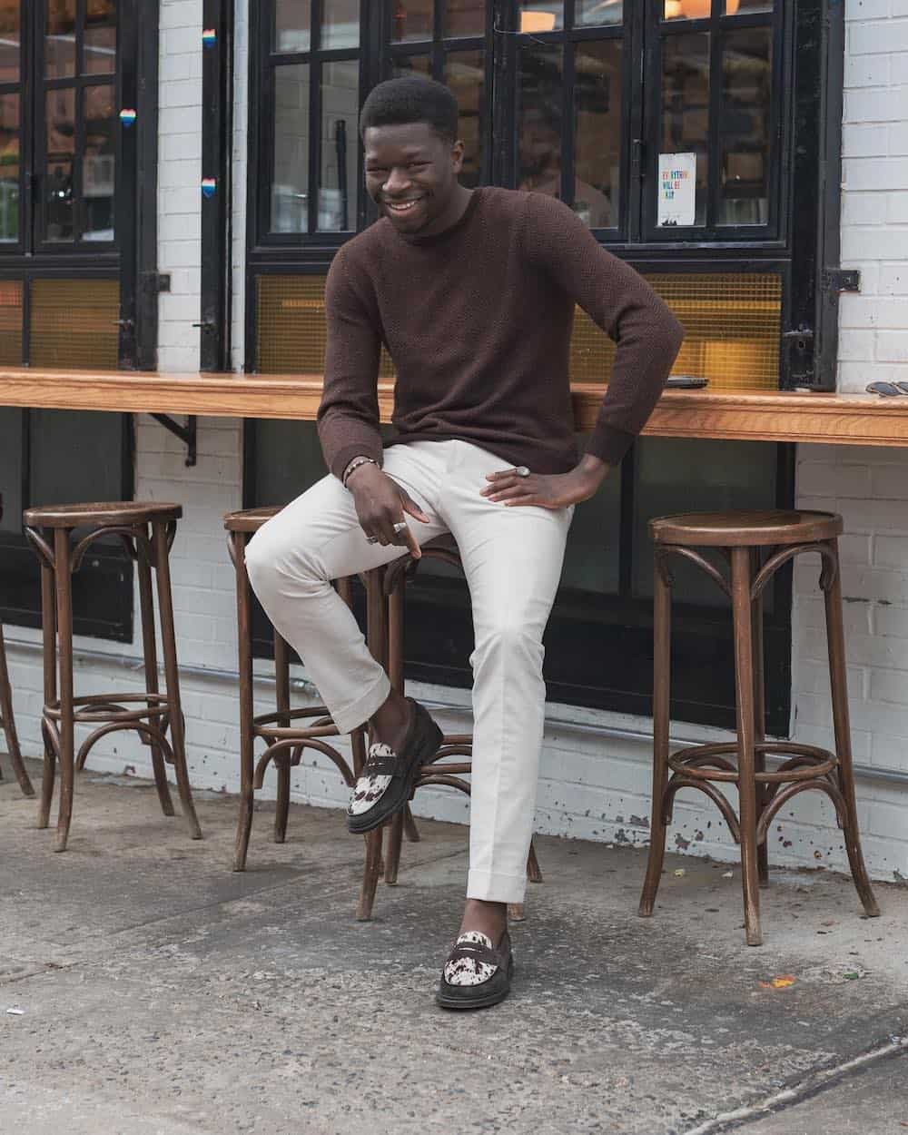 A man wearing white chinos and a brown fine knit sweater with black and white loafers with brown speckles