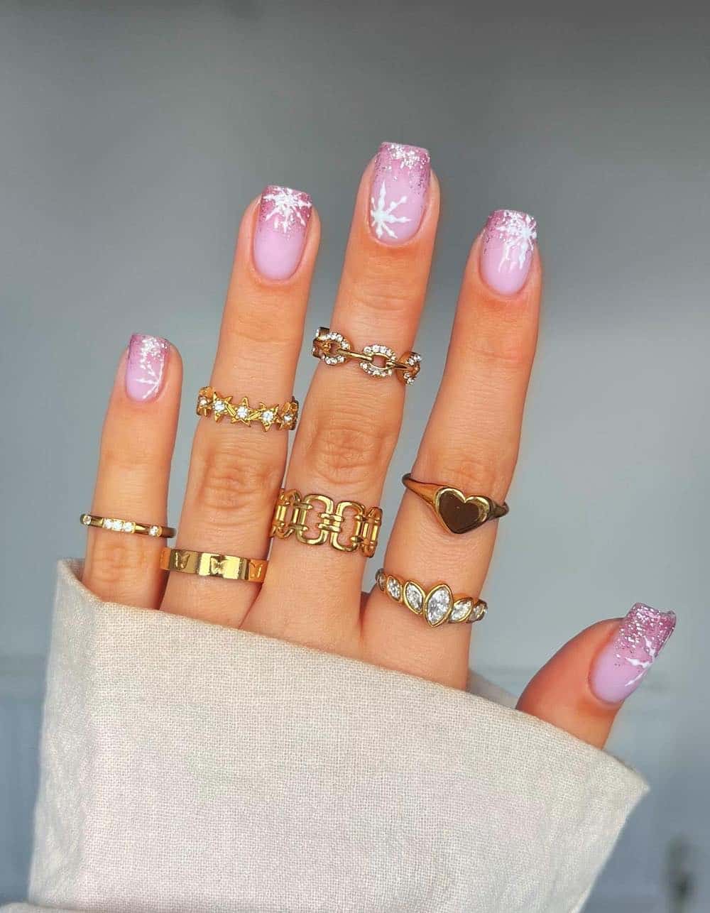 A hand with short nude square nails with pink glitter ombre French tips and white snowflakes