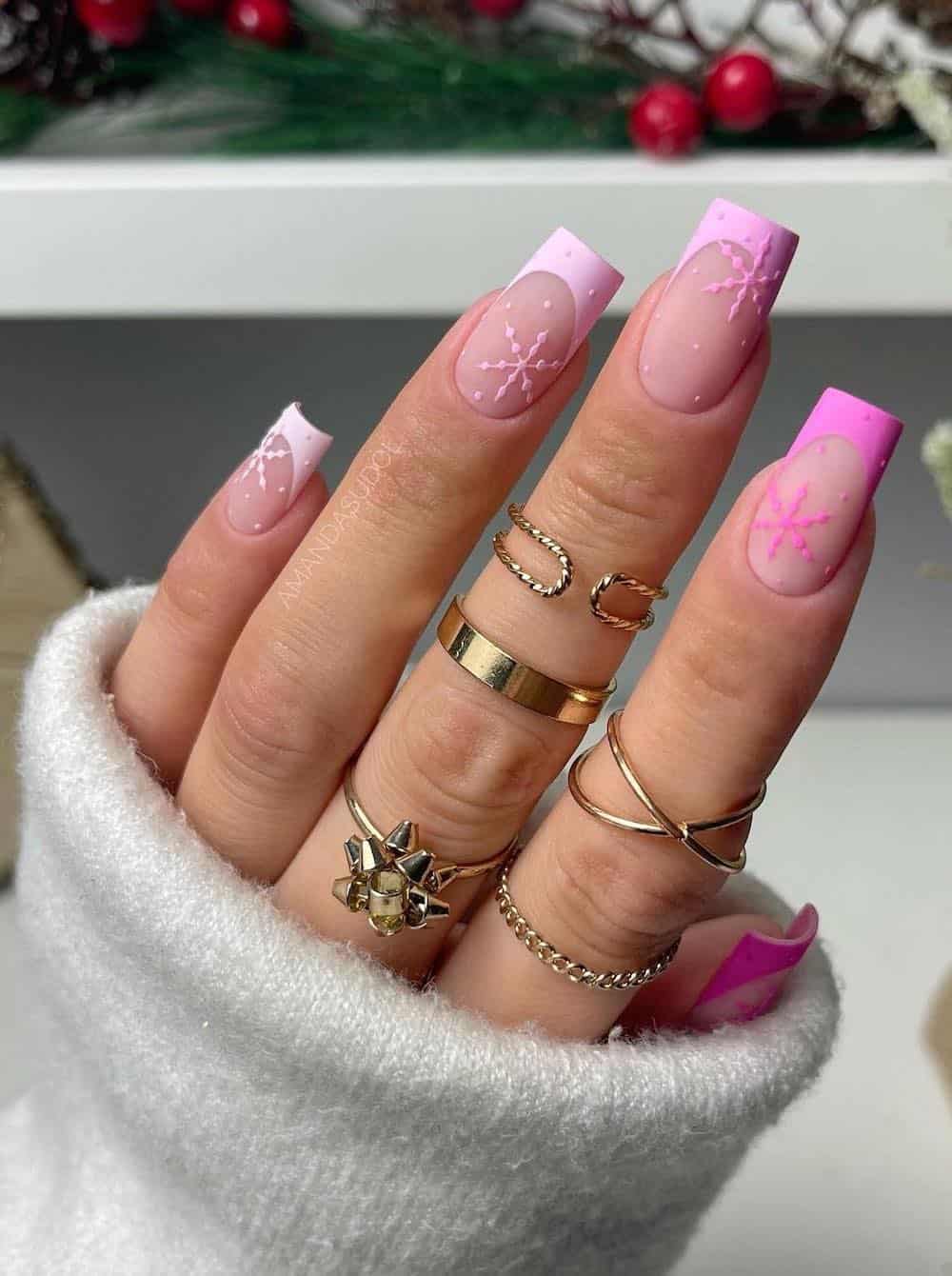 A hand with long square nails with a matte nude base and light to hot pink gradient French tips with snowflake and dot accents