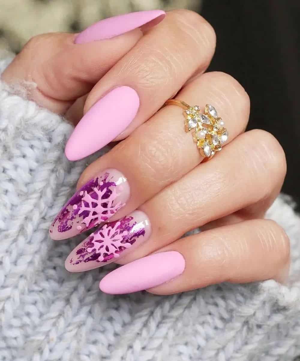 A hand with long light pink almond nails with nude accent nails featuring metallic magenta flakes and light pink snowflakes