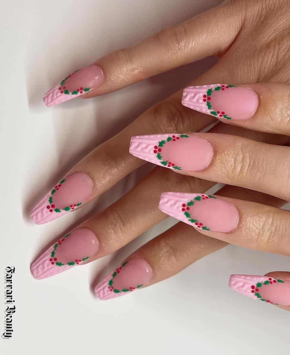 A hand with long coffin nails painted a matte pinkish nude with light pink sweater French tips bordered by wreath and berry details