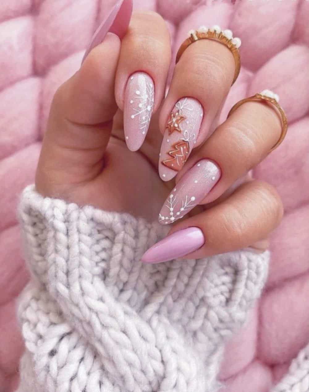A hand with long almond nails painted with glossy light pink polish and sparkly pink polish, featuring white snowflakes and gingerbread cookie nails art