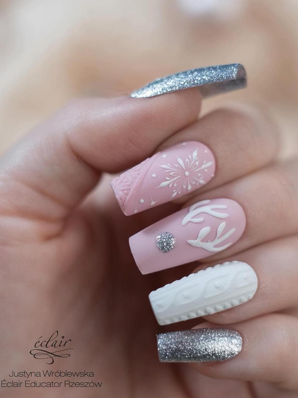 A hand with long square nails featuring silver glitter polish, a white sweater art accent nail, and light pink nails with reindeer and snowflake nail art