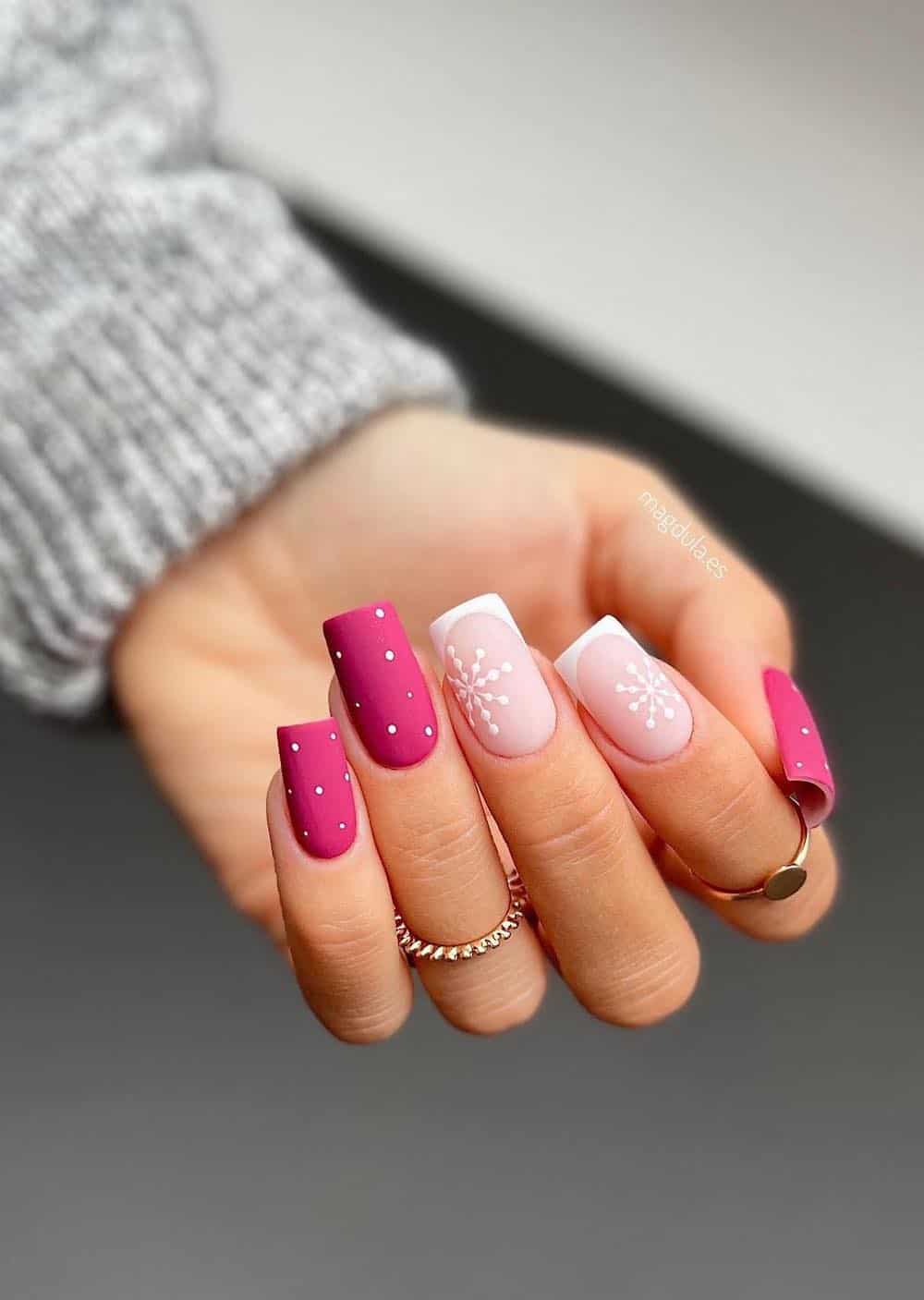 A hand with long square nails painted a matte magenta with white dot details and white French tip accent nails with snowflakes