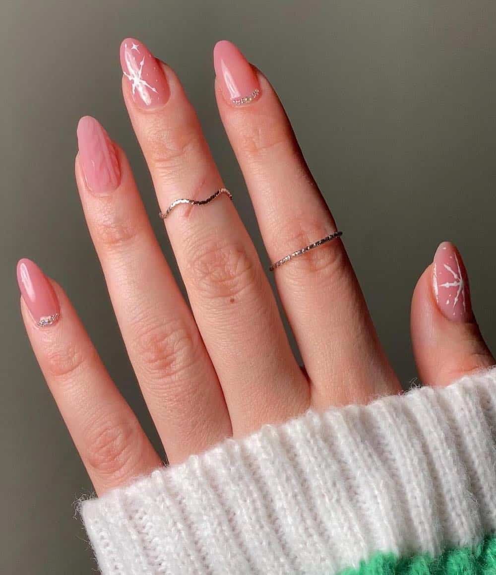A hand with short almond nails painted a glossy pink with silver bead details, white snowflake art, and a matte sweater detail accent nail