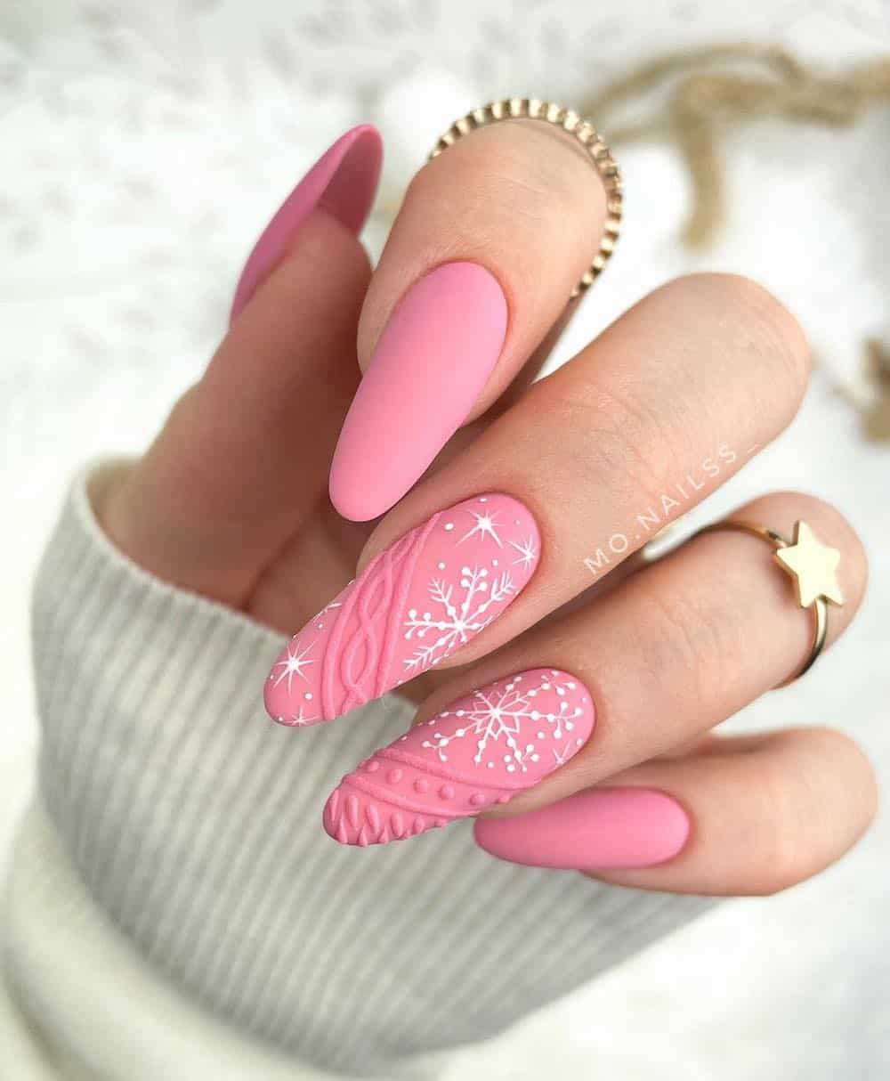 A hand with long almond nails painted a matte light pink with two accent nails featuring sweater details and white snowflakes