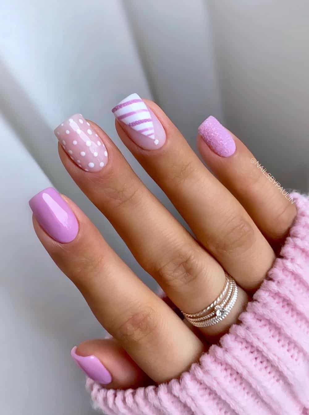 A hand with short square nails painted a glossy pink with accent nails featuring snowy pink polish, nude nails with white dots, and a striped pink and white Santa hat
