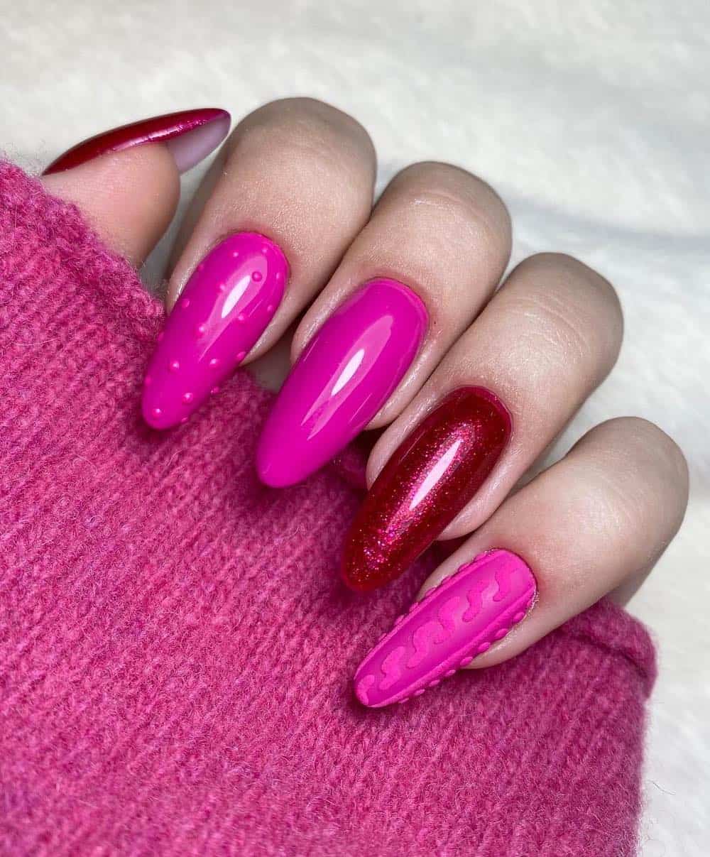 A hand with long almond nails painted a bright pink with dot details and sweater nail art, and two sparkly red accent nails