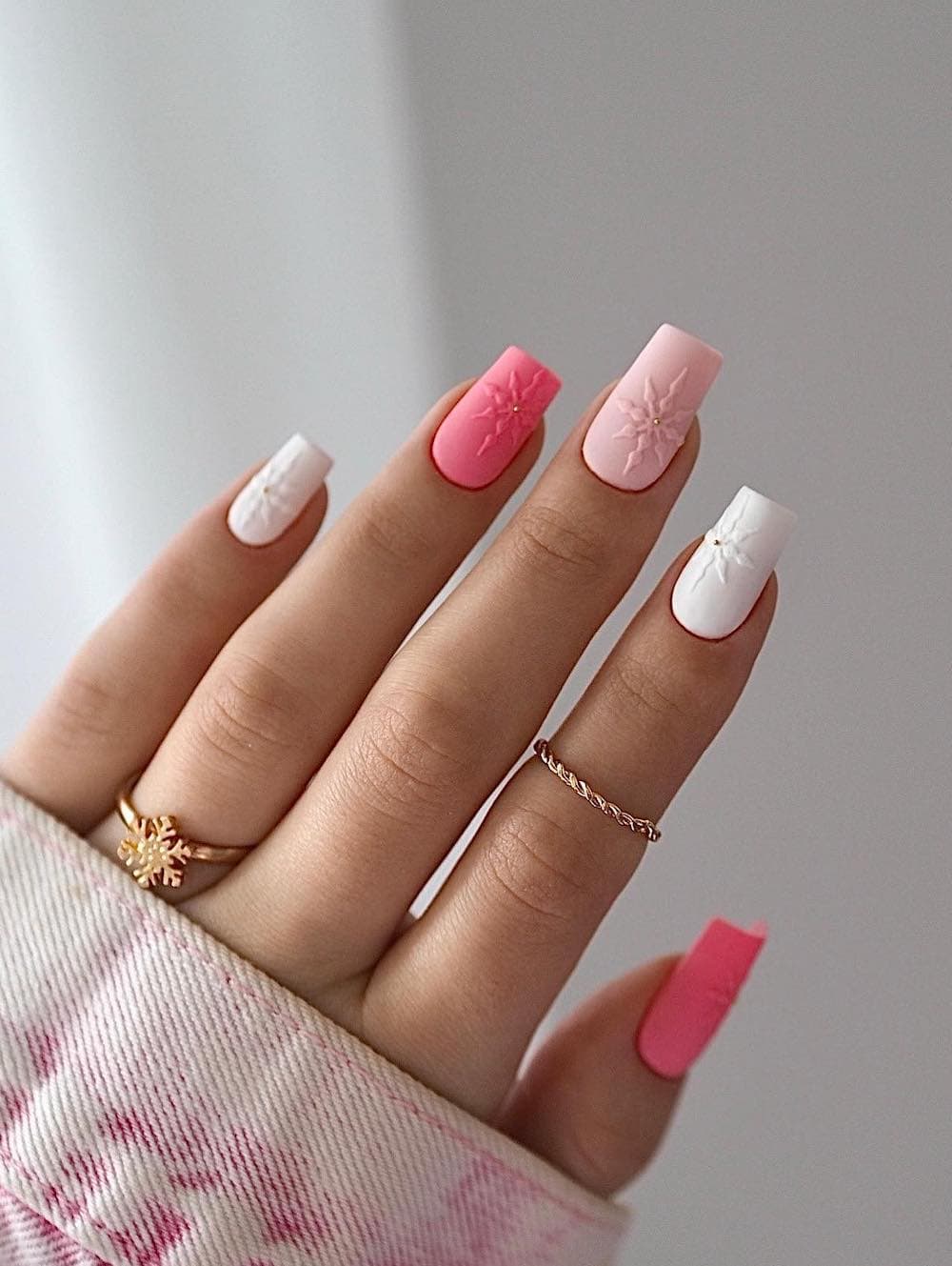 A hand with medium square nails painted in white, light pink, and bright pink with snowflake art and a matte finish