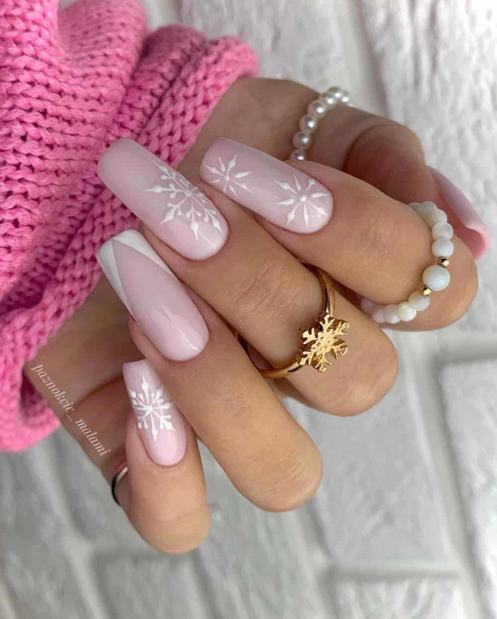 A hand with long square nails painted a soft pink with white snowflakes and a slanted white French tips accent nail