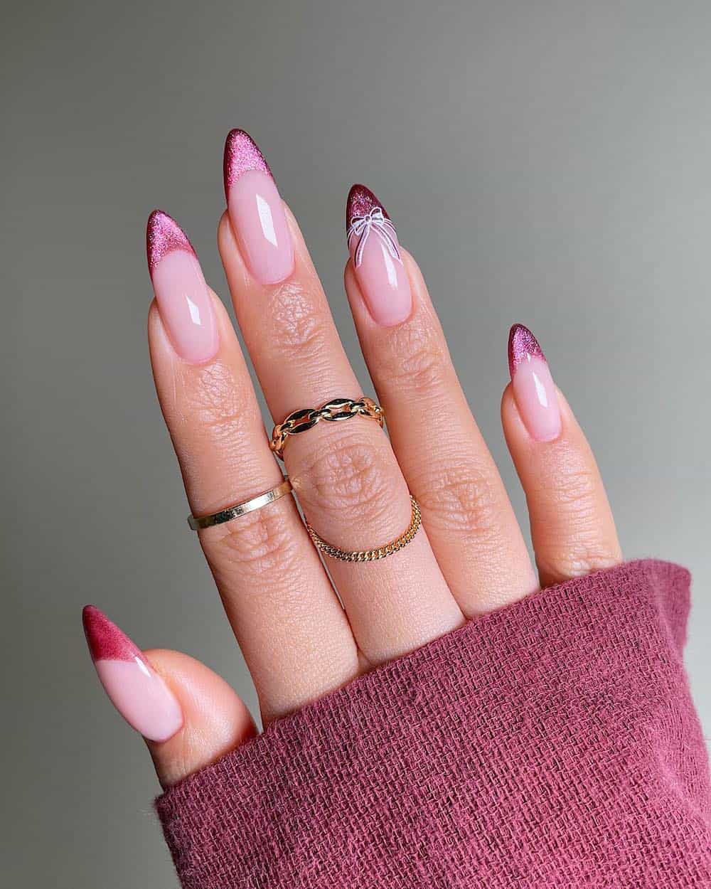 A hand with long glossy nude almond nails with shimmering pink French tips and a bow accent detail