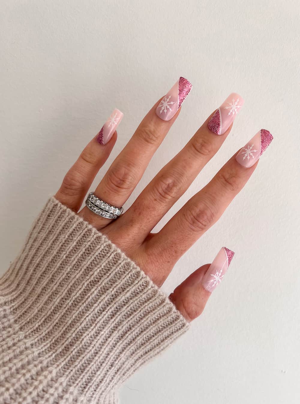 A hand with long square nails painted a nude pink with slanted pink glitter and white snowflakes