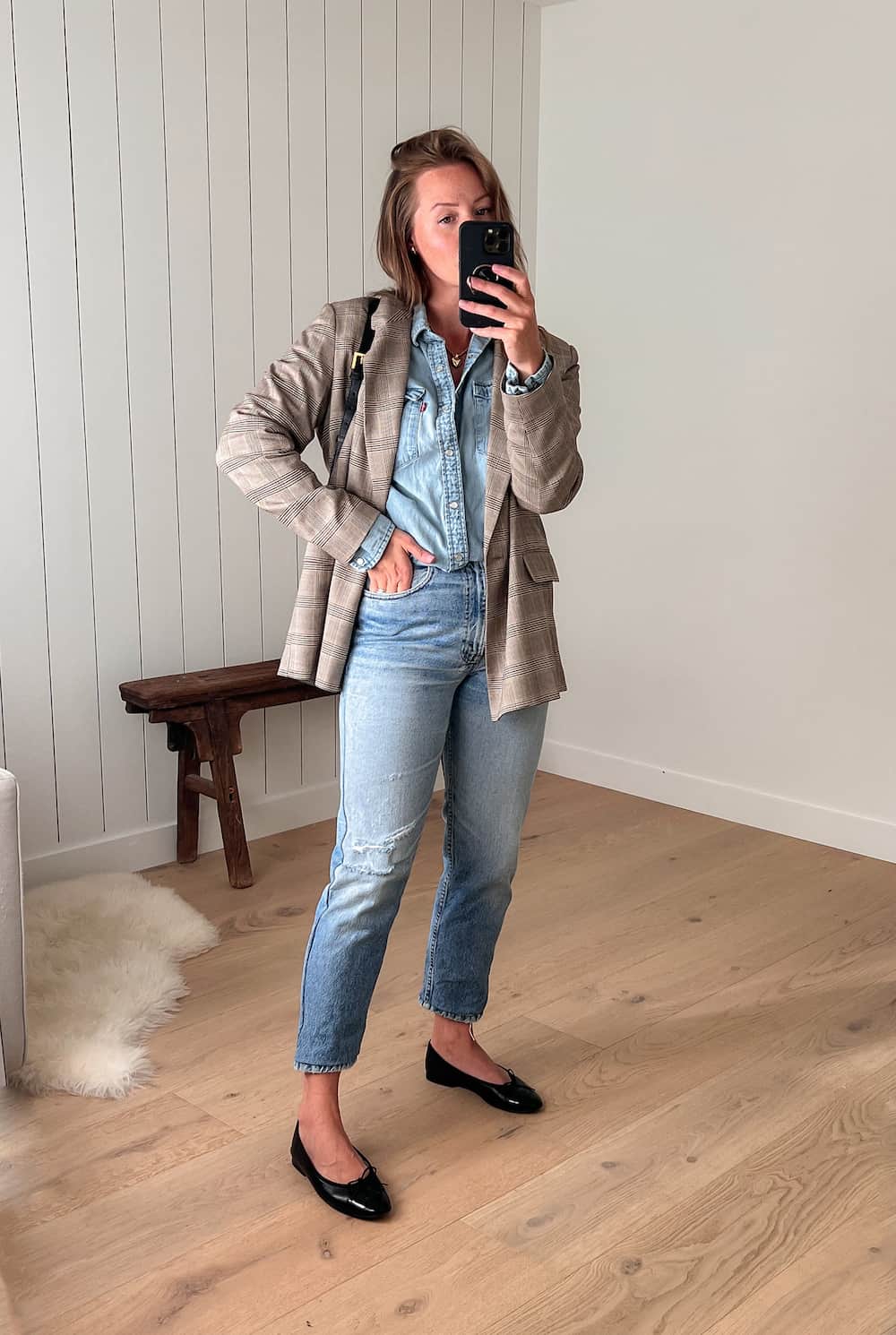 Woman wearing light wash straight leg jeans, a light wash denim button up shirt, a tan plaid blazer and black ballet flats.