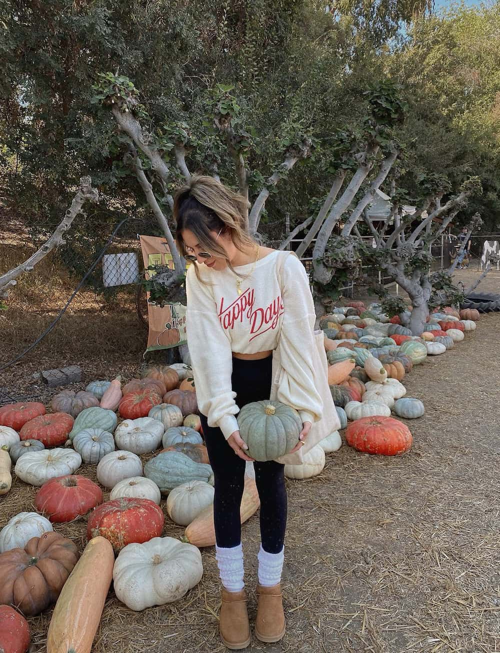 Woman wearing black leggings with a cropped sweatshirt and ugg boots in a pumpkin patch.
