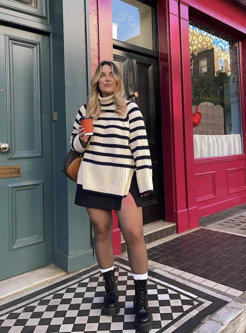Woman wearing a black mini skirt with a black and white striped sweater and black boots outside of a coffee shop.