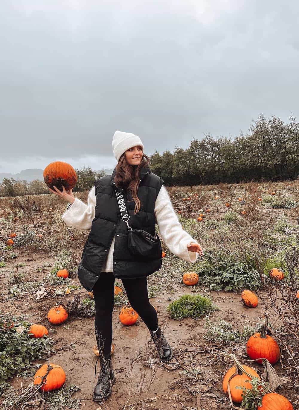 Woman wearing black leggings, black boots, a white sweater, a black puffer vest and a white beanie hat in a pumpkin patch.