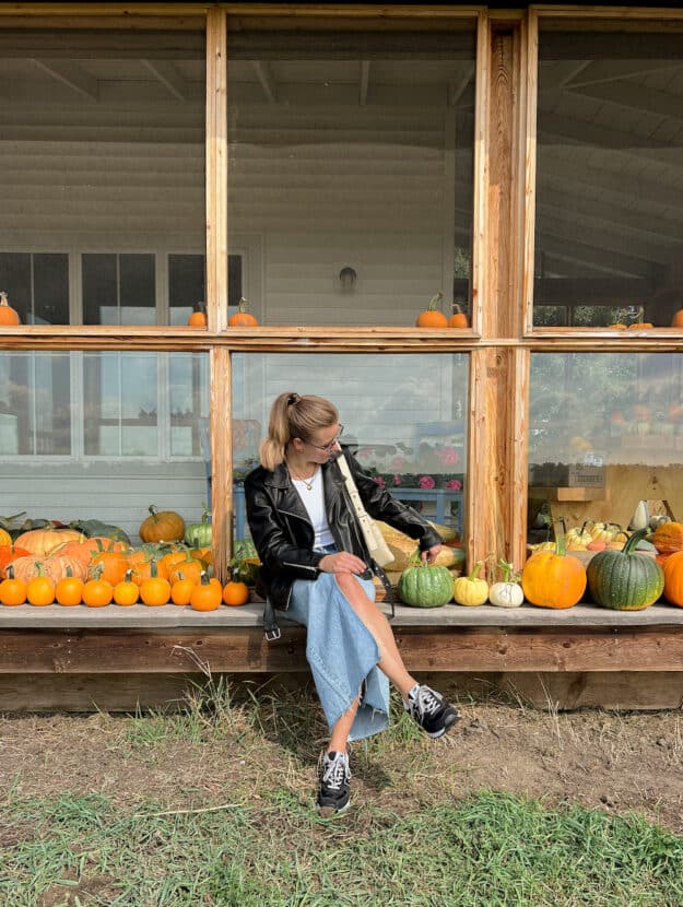Woman wearing a denim maxi skirt, black sneakers, a white t-shirt and a black leather jacket in a pumpkin patch.