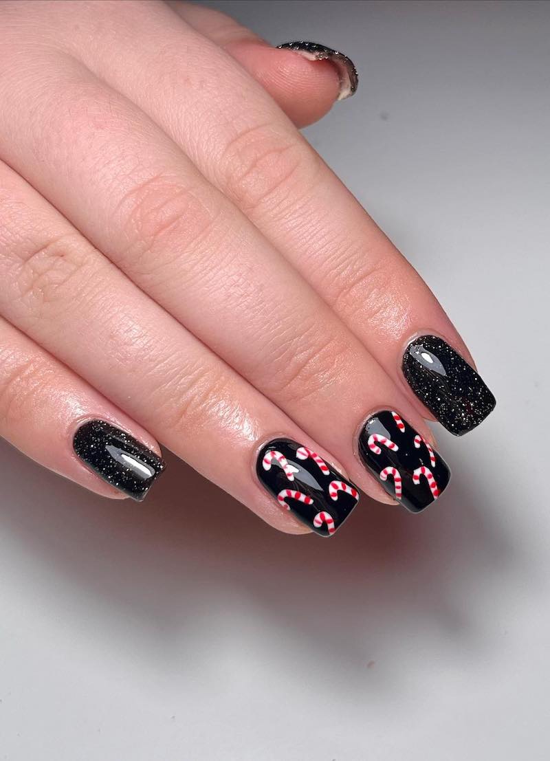 A hand with short black nails with white speckles and two accent nails with candy cane nail art