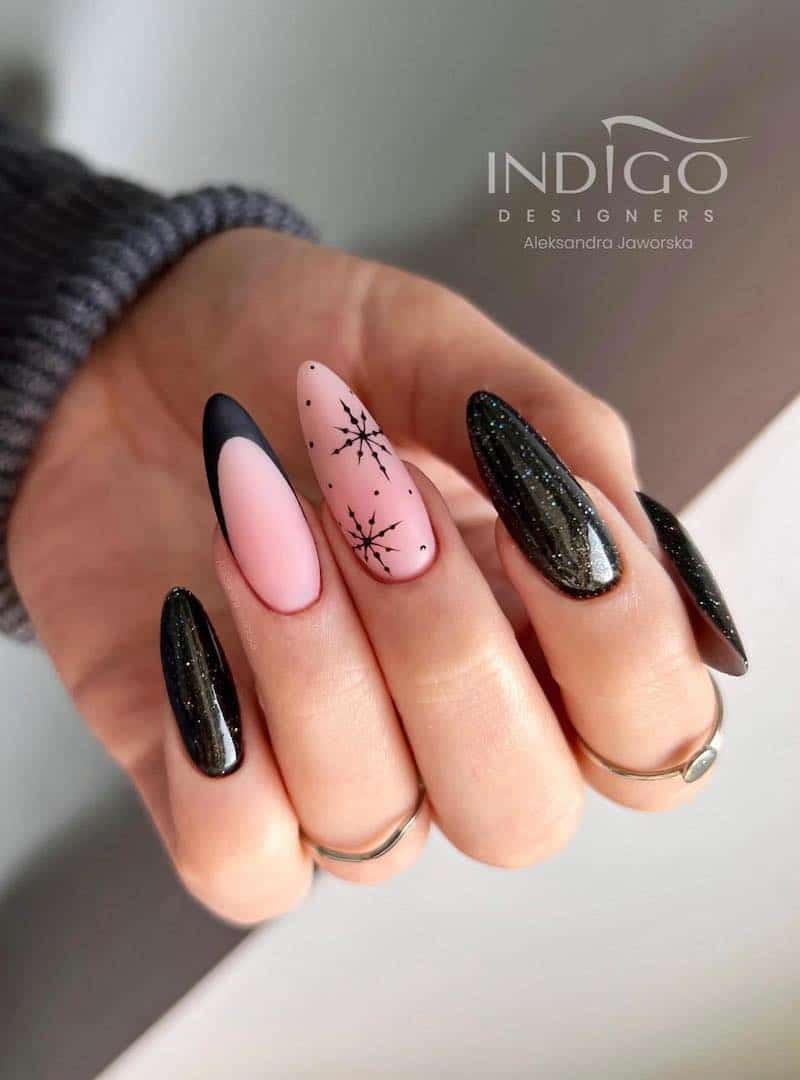 A hand with long almond nails painted with glittering black polish and two nude pink accent nails with a black French tip and black snowflakes