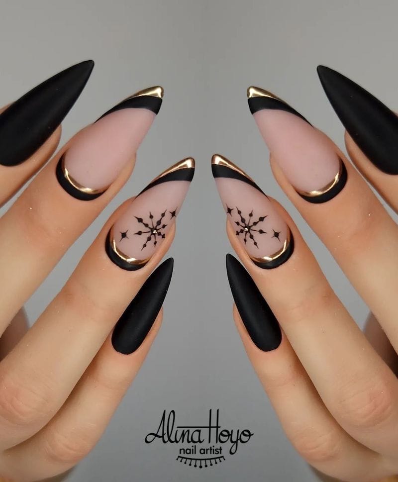A hand with long black stiletto nails and two matte nude accent nails with black and gold borders and black snowflakes