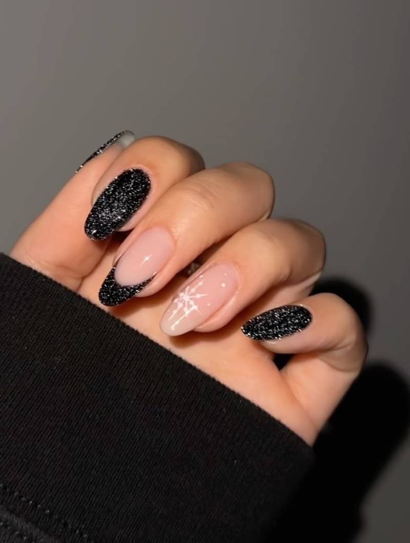 A hand with medium almond nails painted with glittery black polish with a French tip accent nail and a nude accent nail with a white snowflake