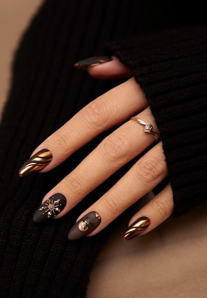 A hand with medium almond nails painted a matte black with gold polish waves and two accent nails with gold snowflakes and Christmas ornaments