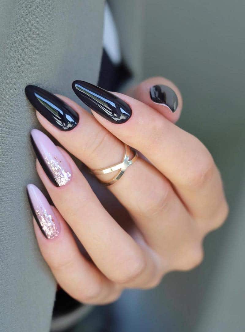 A hand with long almond nails painted a glossy black with two accent nails featuring nude pink and black with gold glitter