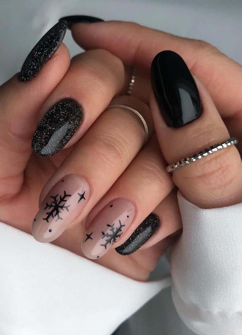 A hand with medium round nails painted a glittering black polish, two nude accent nails with black snowflakes, and another hand with plain glossy black nails