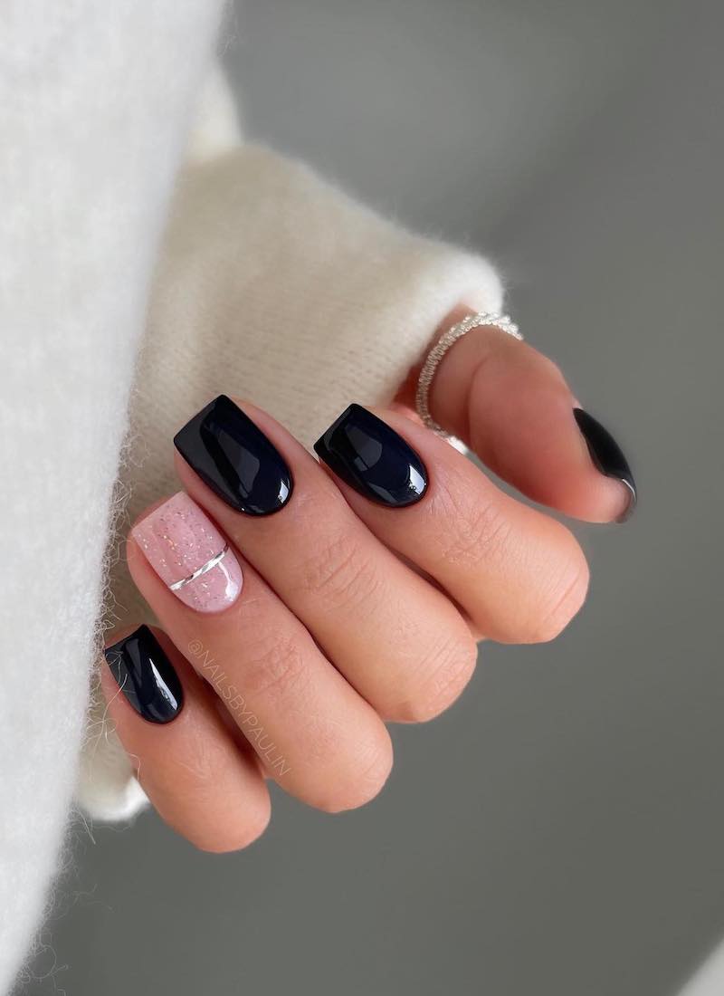 A hand with short square nails painted a glossy black with a nude pink accent nail with glitter and a silver band