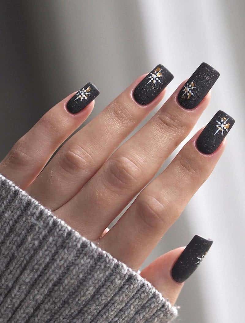 A hand with long square nails painted a matte black with white-speckled polish, white snowflakes, and gold accents