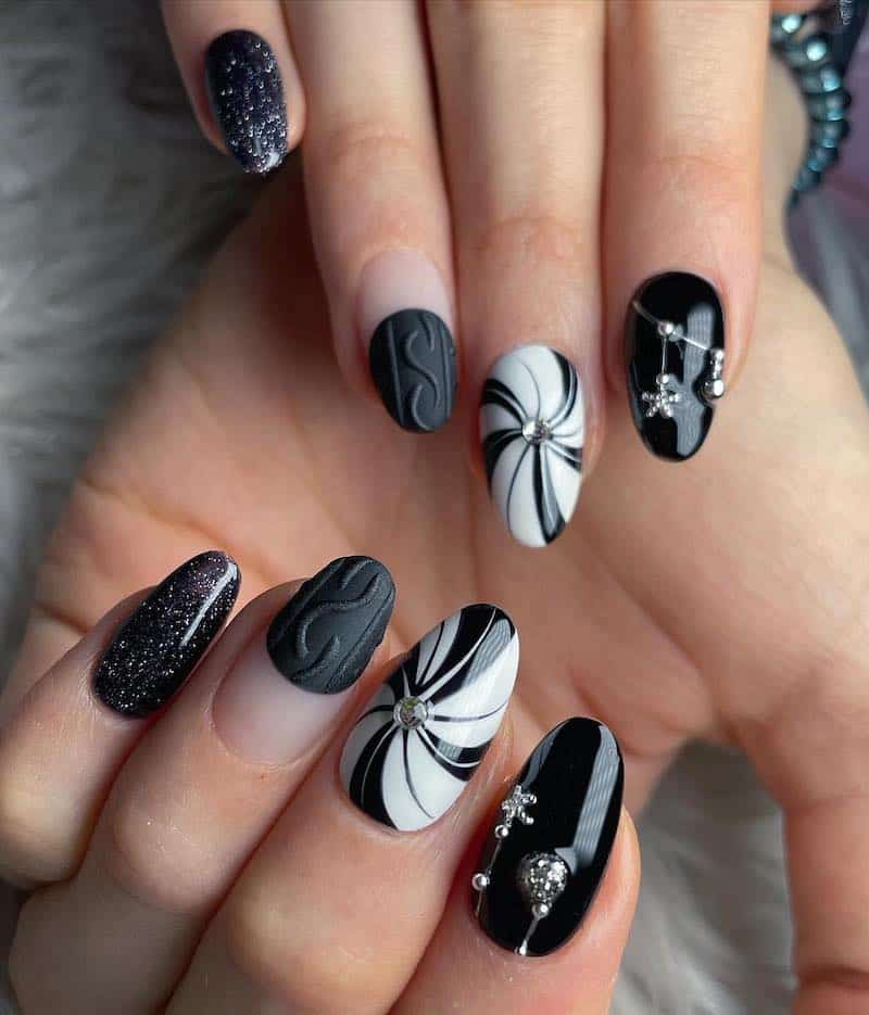 A hand with medium almond nails with multiple designs including black polish with silver ornament beads, matte sweater French tips, glitter nails, and black and white peppermint swirl nails