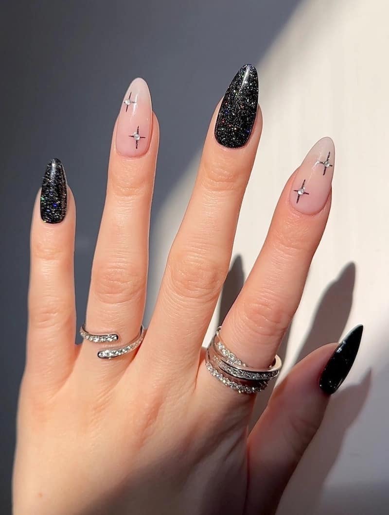 A hand with medium almond nails painted a glittering black with two nude accent nails featuring sparkles with gems