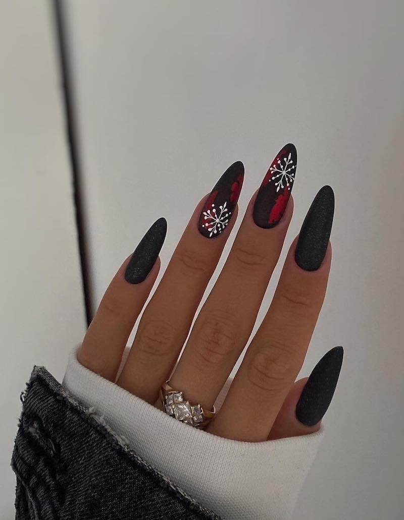 A hand with long almond nails painted a glittering black with two black accent nails featuring metallic red details and white snowflakes