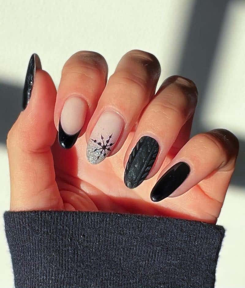 A hand with short almond nails painted with black polish featuring a silver glitter ombre nail with a black snowflake, black French tips, and a sweater nail