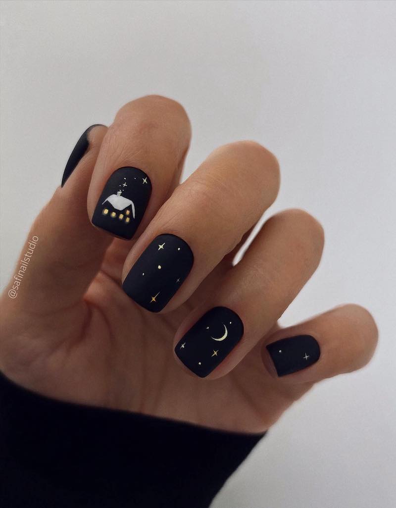 A hand with short squoval nails painted a matte black with star and moon art and an accent nail with a snowy house painted on it
