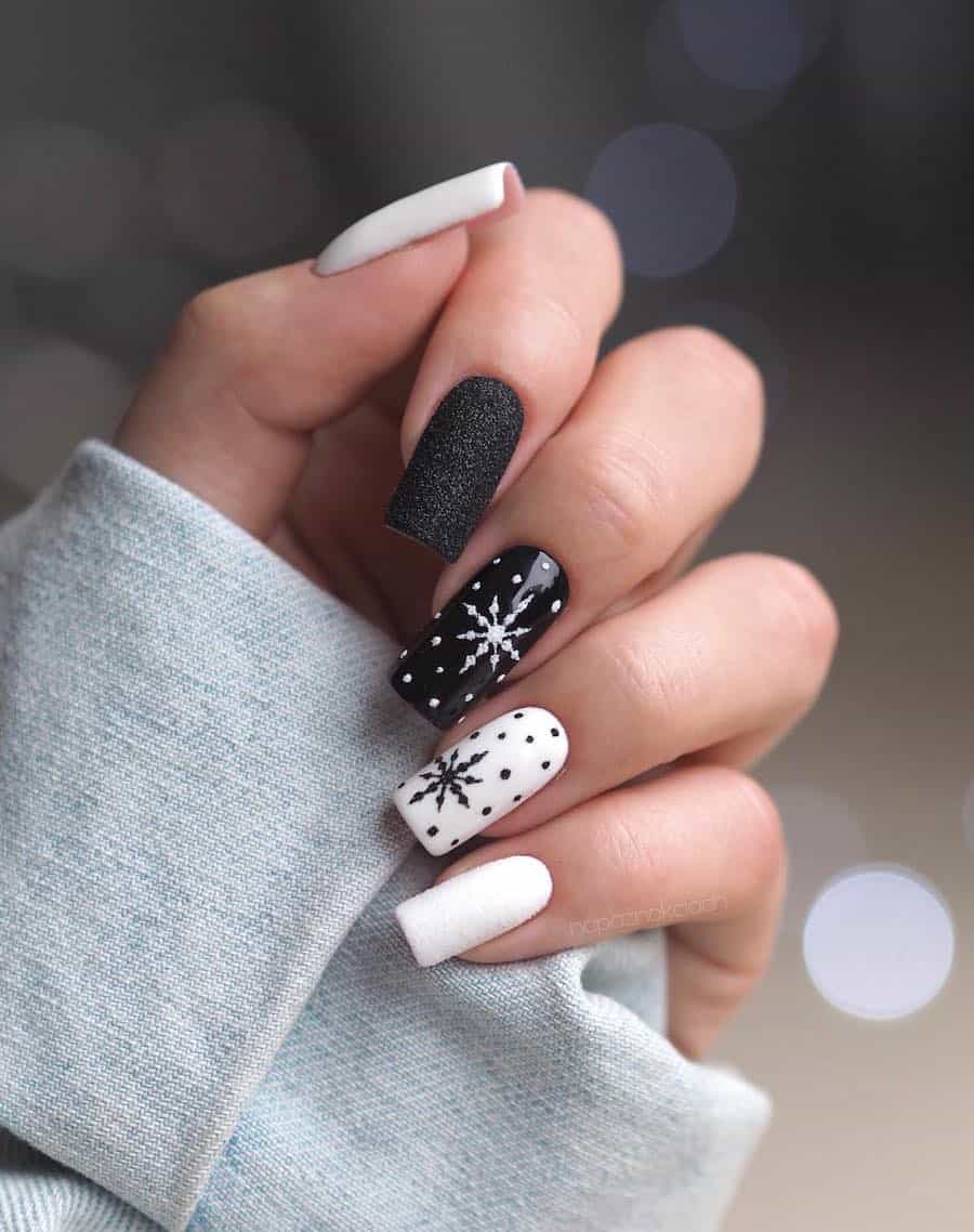 A hand with long square nails painted white and black with snowy texture and two glossy black and white nails with dots and snowflakes