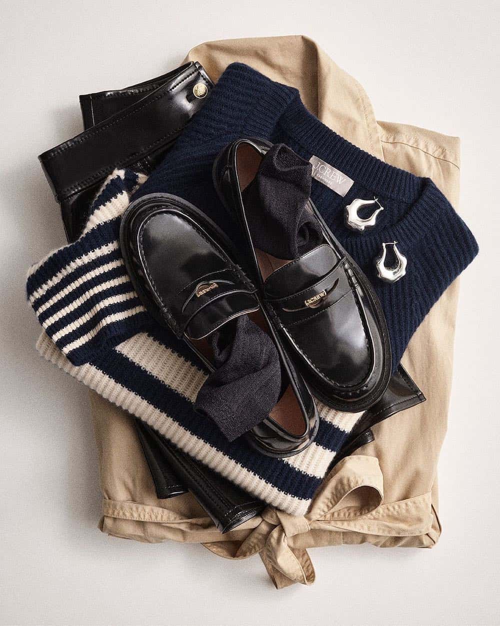 image of a stacked pile of clothing including a trench coat, striped sweater, and black penny loafers from J.Crew 