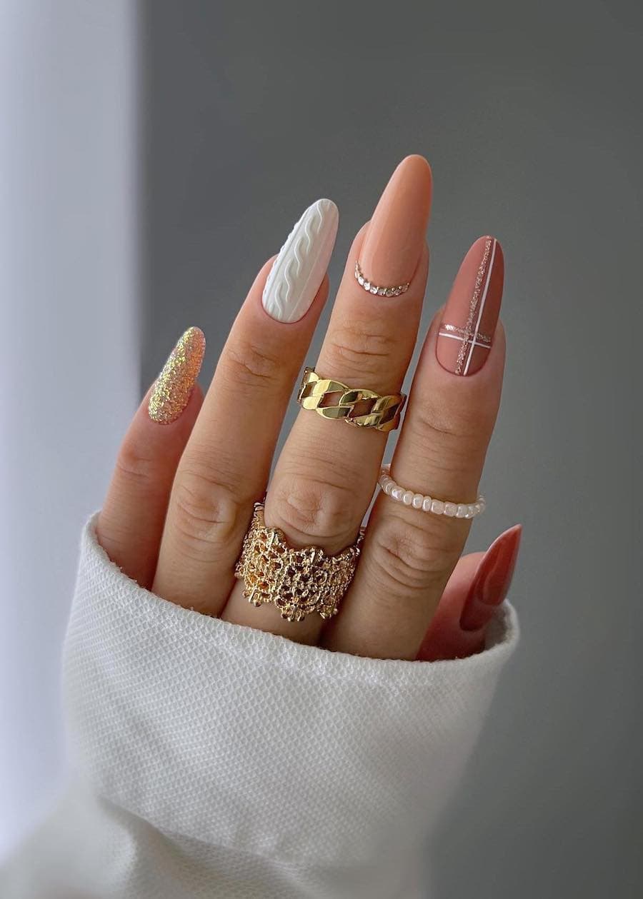 A hand with long almond nails painted in multiple designs, including gold glitter, white sweater details, glossy beige with gem details