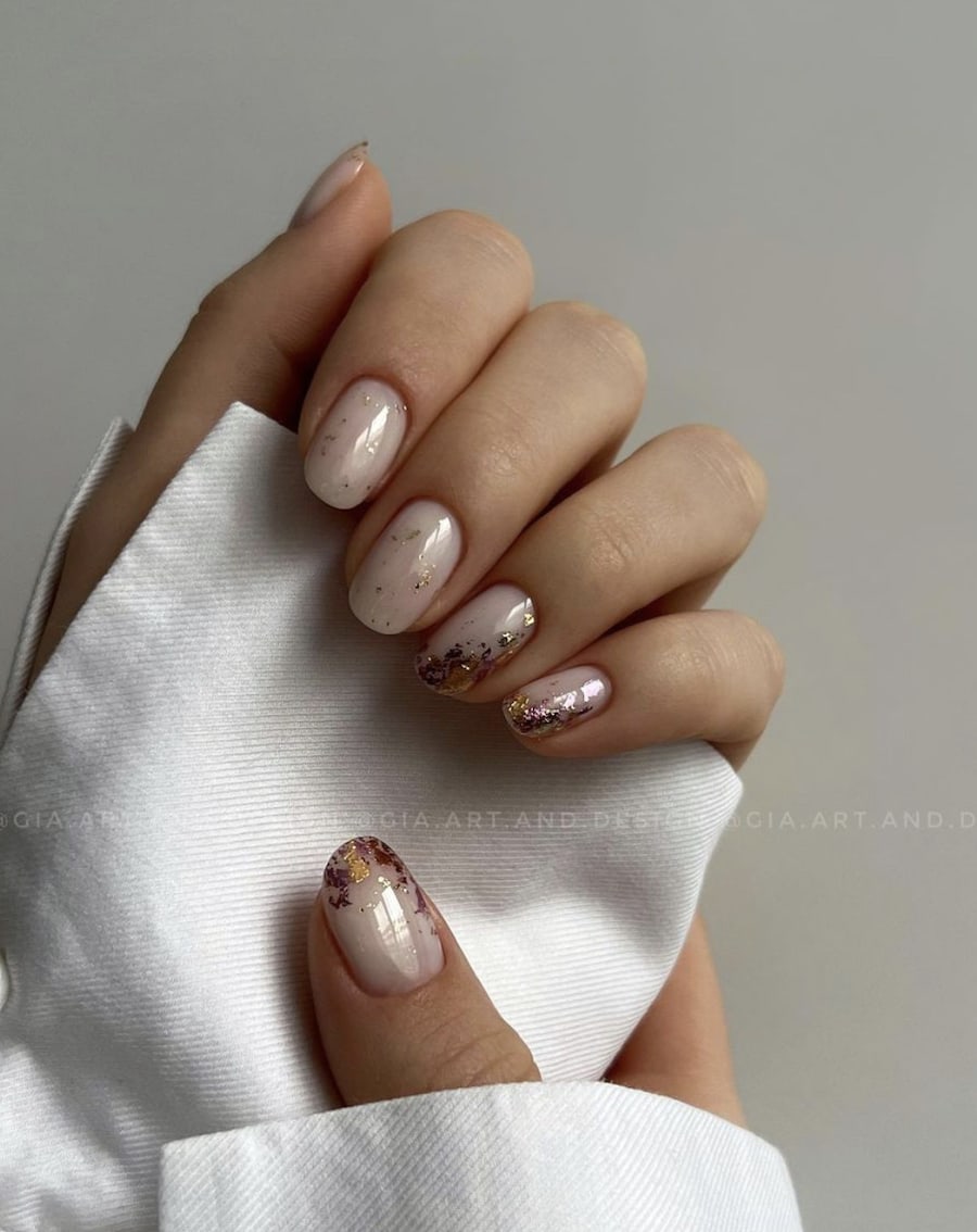 A hand with short round nails painted a milky white with gold and pink metallic flakes