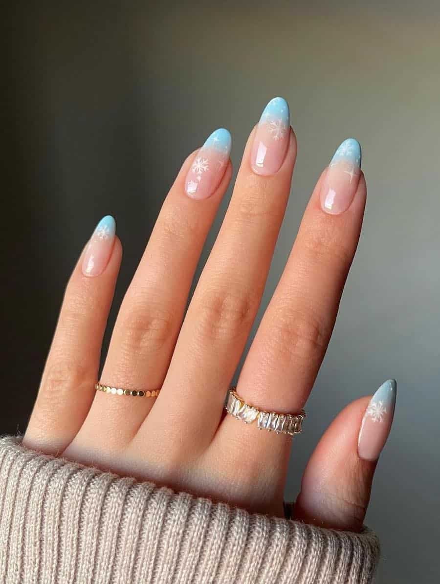 A hand with medium almond nails painted a light blue and nude ombre with white snowflakes