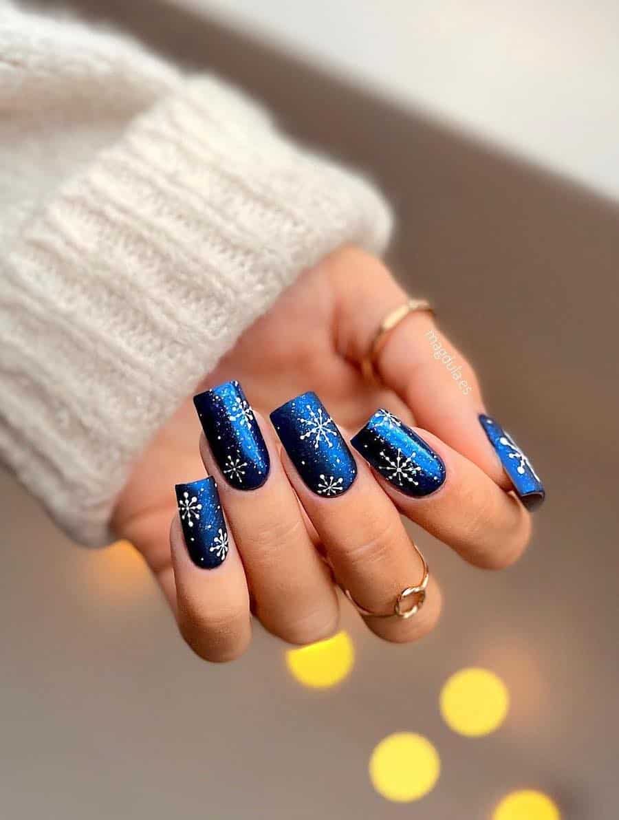 A hand with long square nails painted a shimmering dark blue with white splatter accents and snowflakes