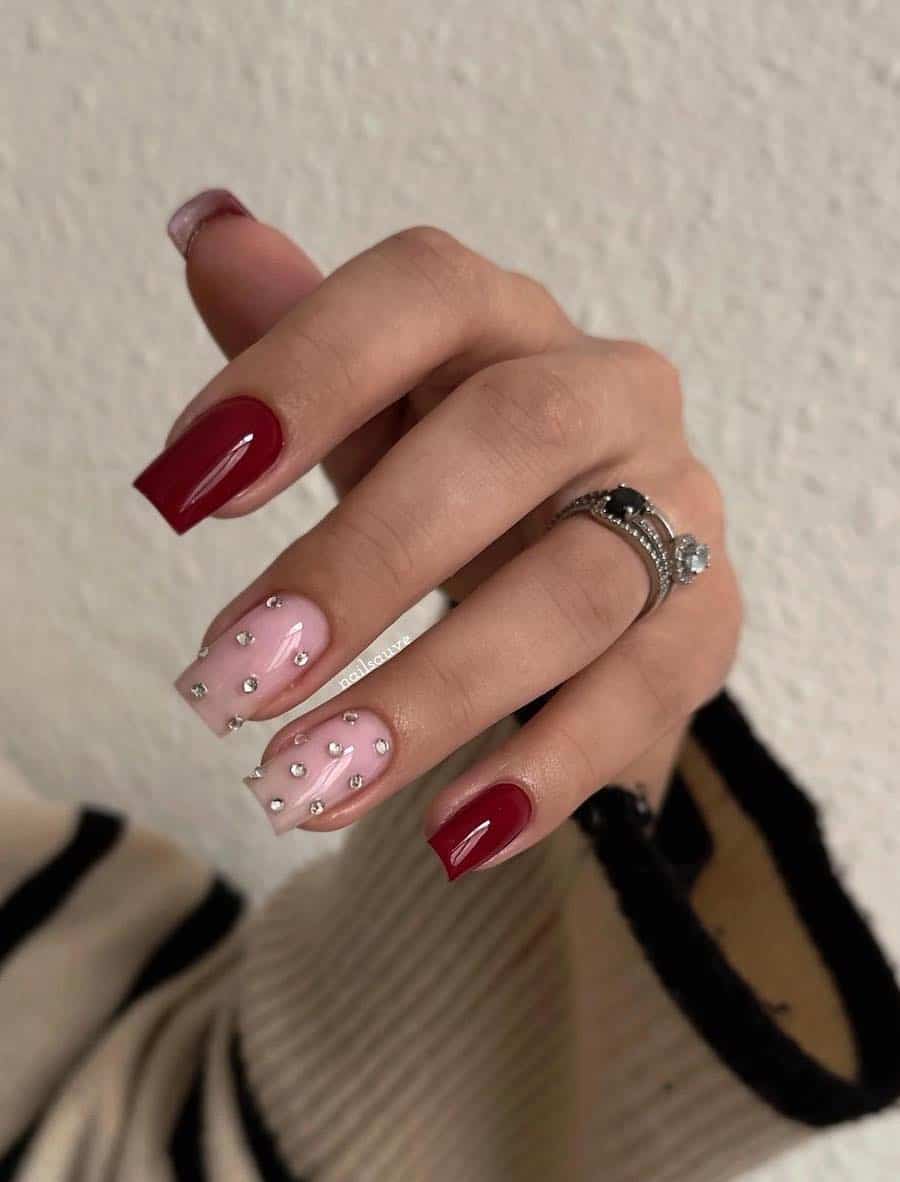 A hand with short square nails painted a dark red with two glossy nude pink accent nails with crystals