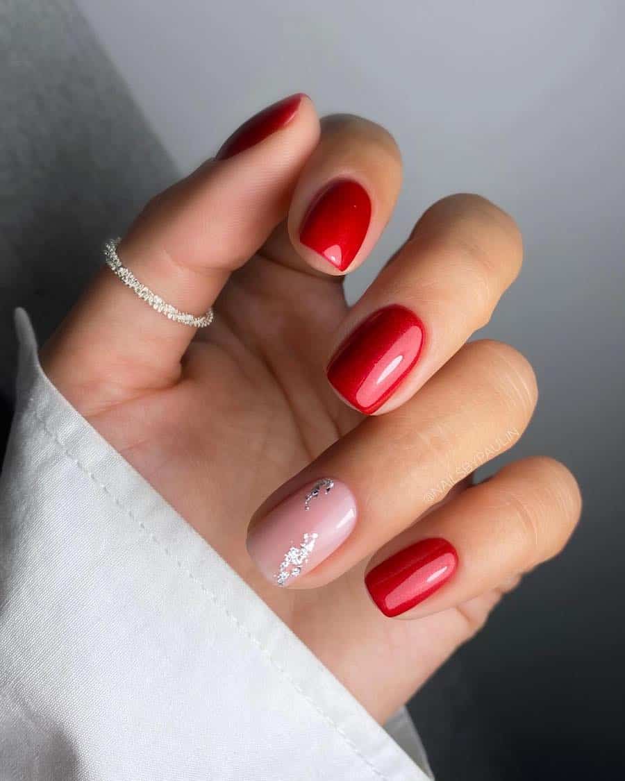 A hand with short squoval nails painted in red polish with one nude accent nail with silver flakes