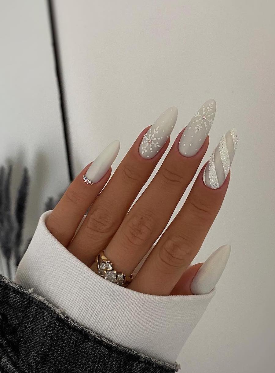 A hand with long white almond nails painted with multiple designs, including shimmering white stripes, snowflake art, and crystal accents