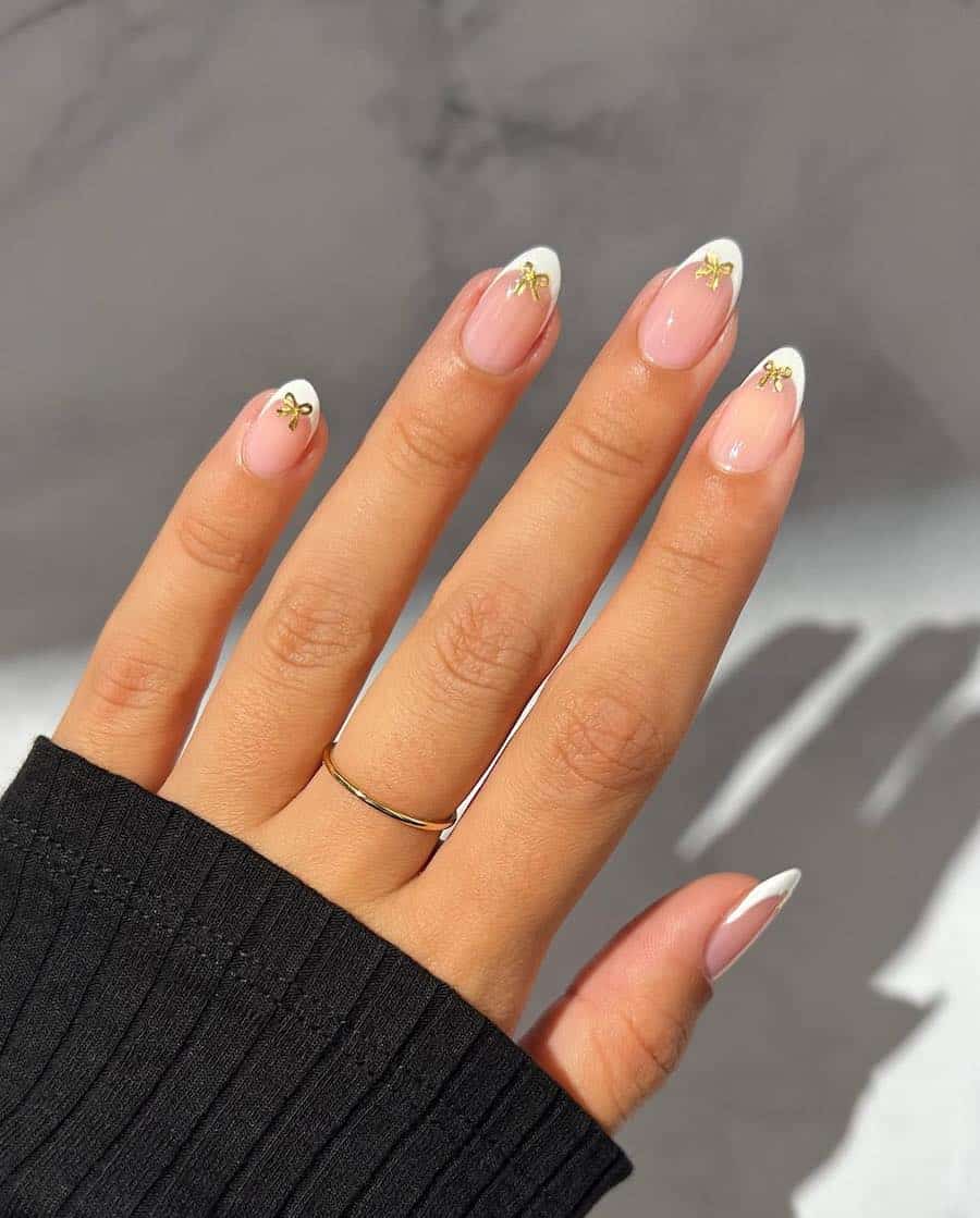 A hand with short almond nails painted with white French tips on nude pink nails with gold bow details