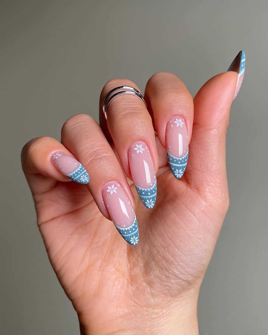 A hand with long almond nails painted a glossy nude pink with blue and white sweater detailed French tips and white snowflake accents