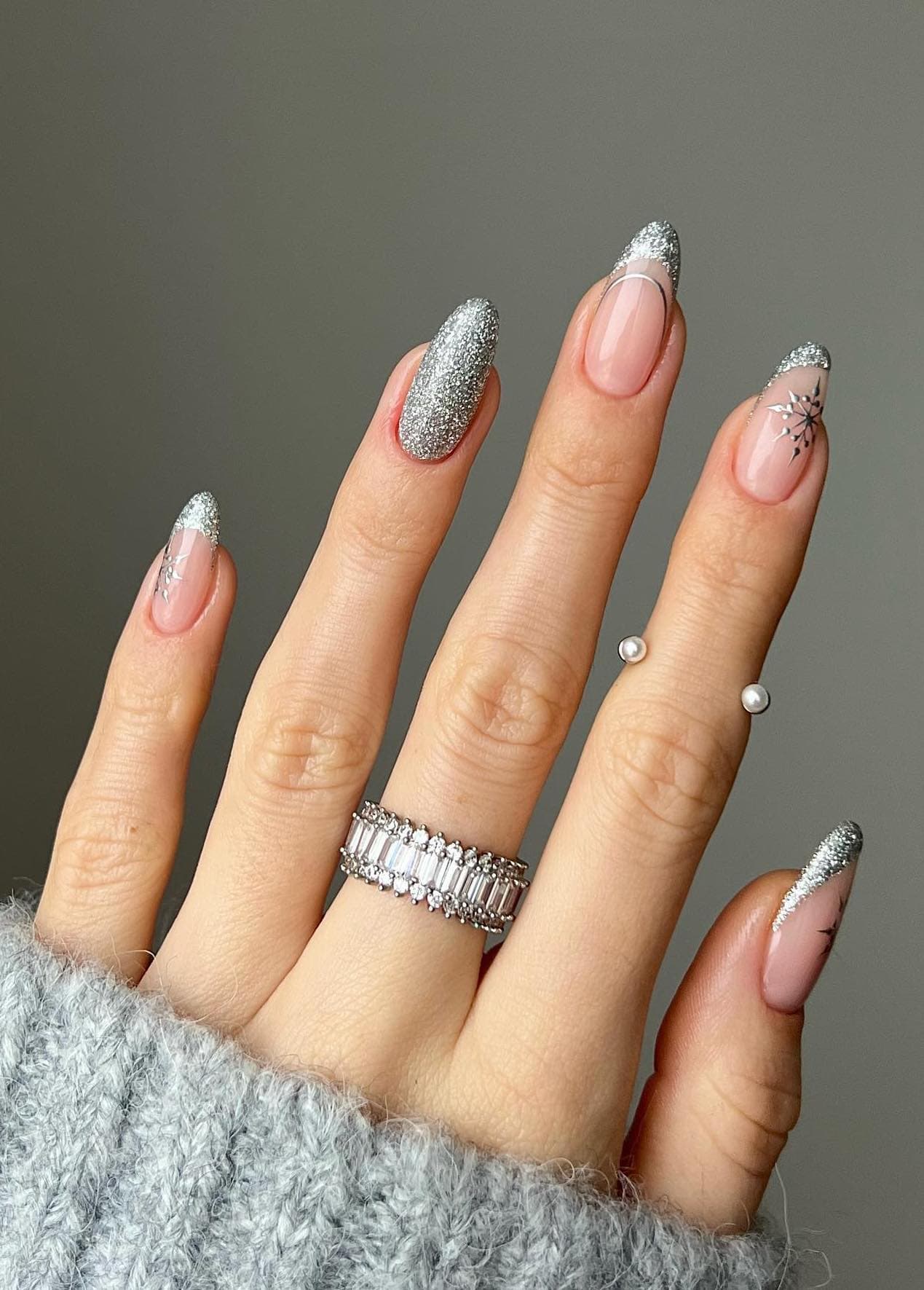 A hand with medium nude almond nails painted with silver glitter French tips with a full silver glitter accent nail and silver snowflake accents