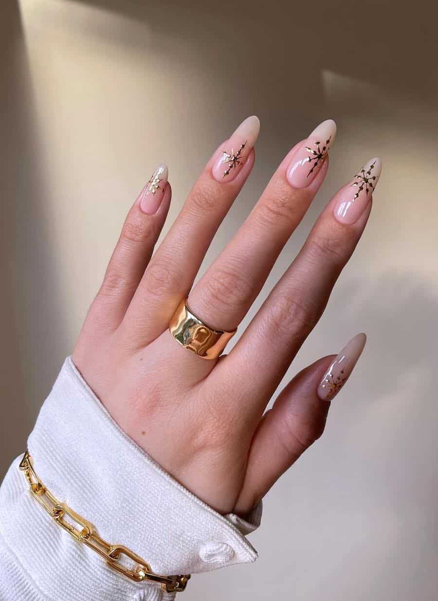 A hand with long nude almond nails with gold snowflakes