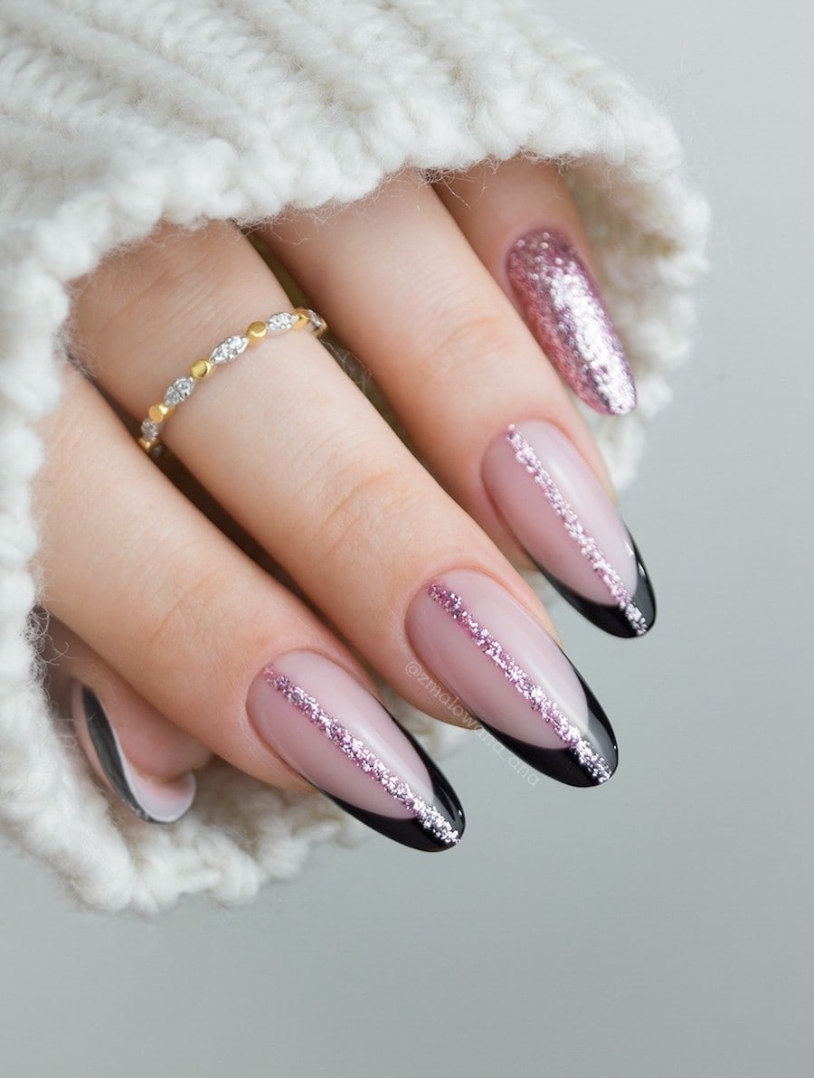 A hand with long nude pink almond nails with black French tips and vertical pink glitter lines down the center of each nail