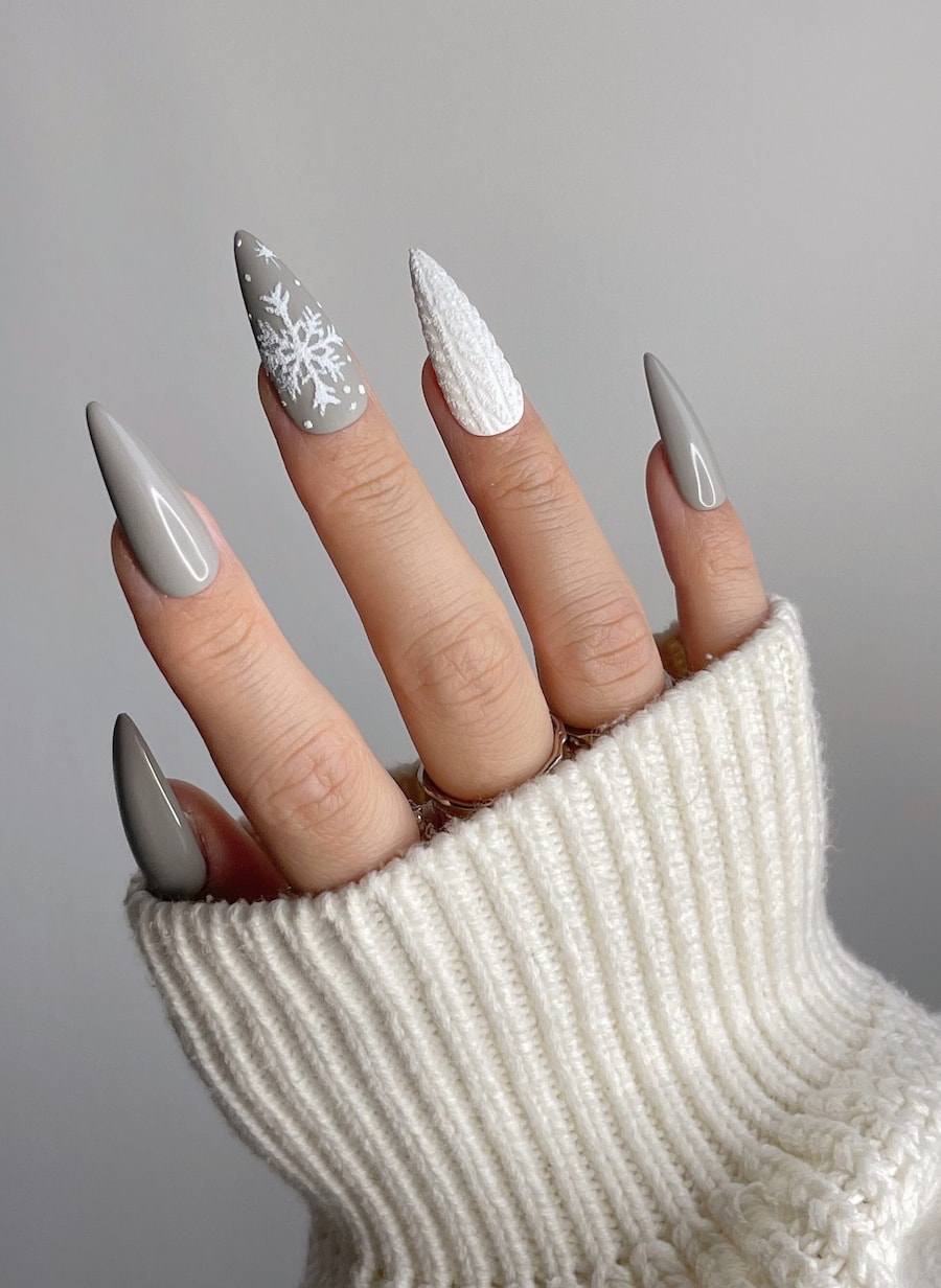 A hand with long stiletto nails painted a glossy grey color with a white sweater accent nail and a matte grey nail with white snowflakes